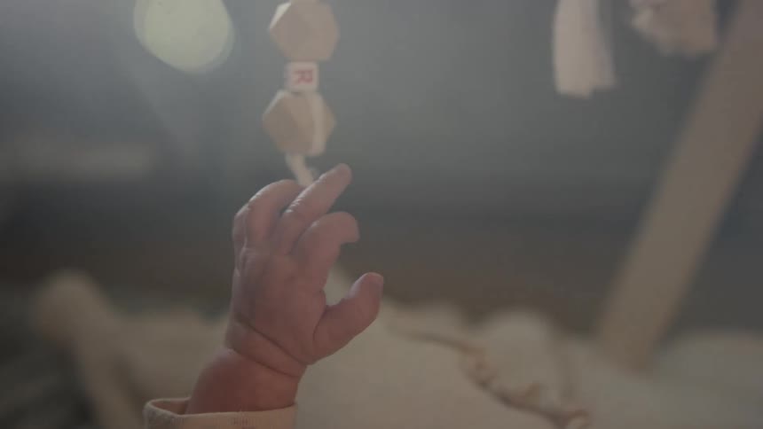 An intimate slow-motion close-up captures a newborn baby's tiny hand reaching for a hanging mobile in a softly lit nursery