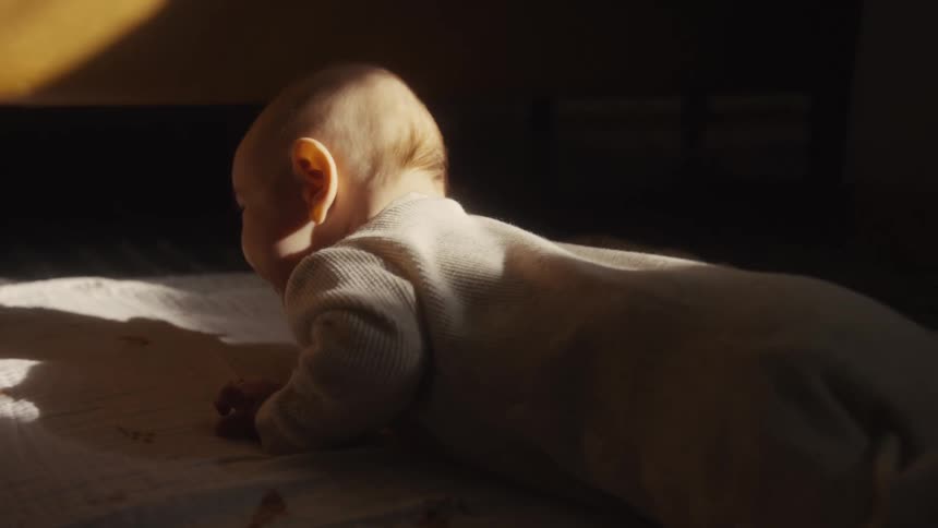In a quiet, dark room, a newborn baby in a white onesie practices tummy time while bathed in a warm, dramatic strip of sunlight