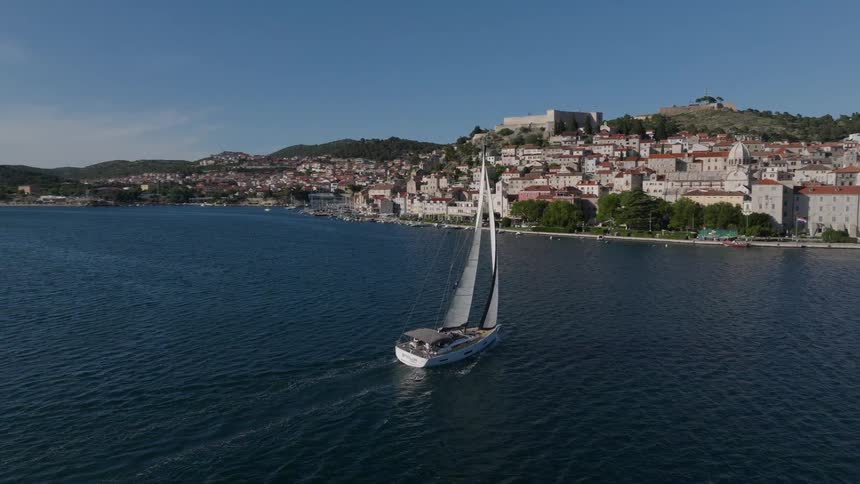 An aerial shot follows a beautiful white sailboat as it glides across the water past a historic European coastal town on a sunny day