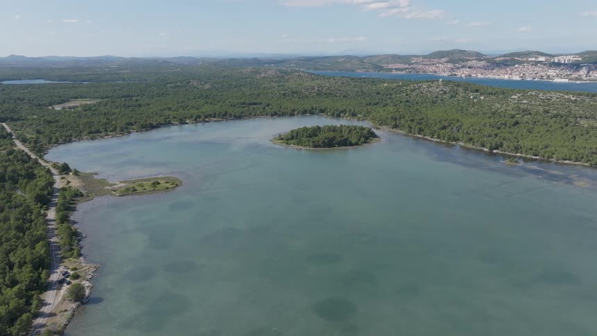 A breathtaking aerial perspective of a tranquil turquoise bay, featuring a lush forested island and a distant coastal city under a clear blue sky