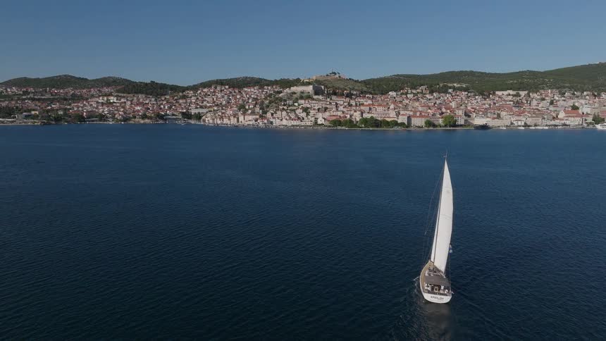 An aerial drone shot follows a sailboat gliding across the deep blue Adriatic Sea, pulling back to reveal the beautiful coastal city of Šibenik, Croatia
