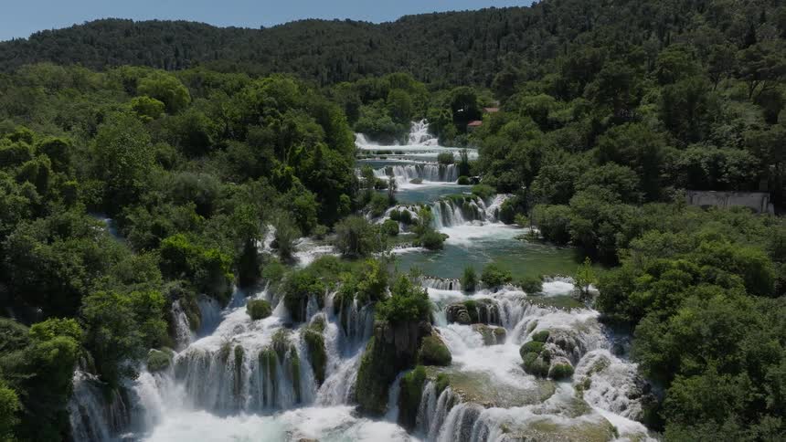 Aerial view slowly pulling back to reveal a majestic multi-tiered waterfall flowing through a lush green forest on a sunny day