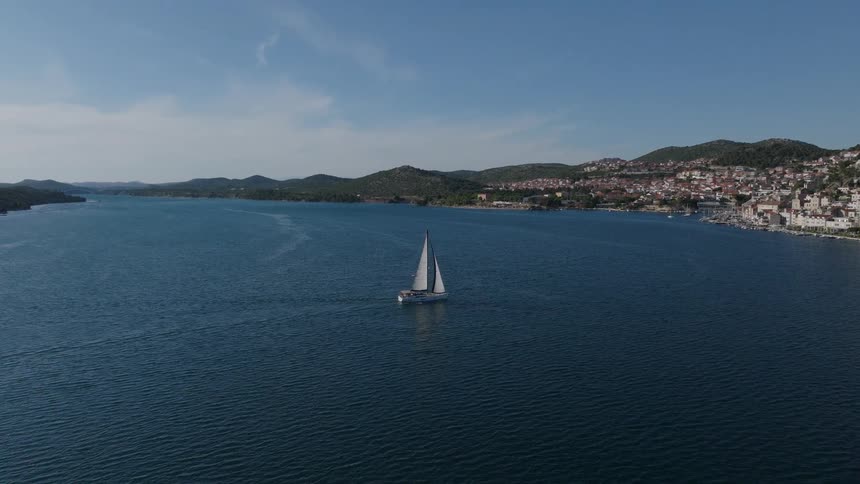 A beautiful aerial view shows a sailboat gliding across the tranquil blue sea towards a scenic coastal town under a clear sky