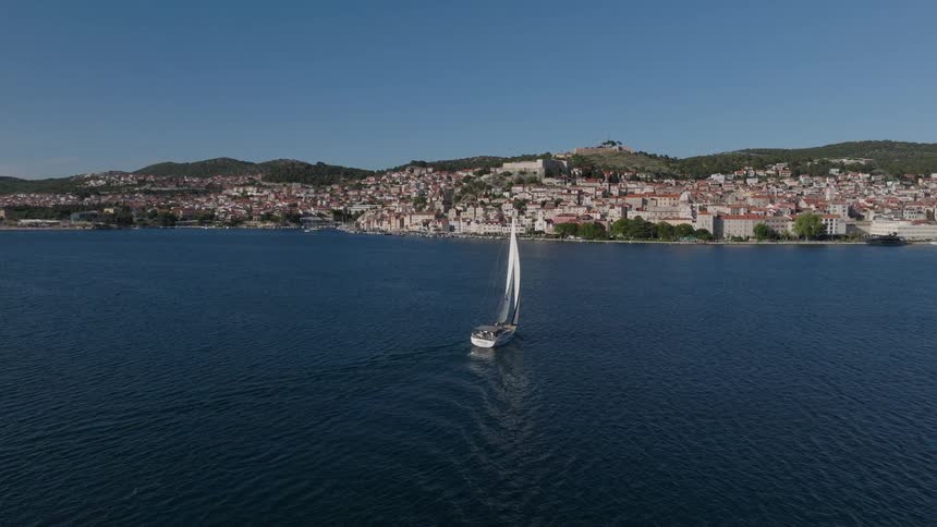 Aerial footage follows a sailboat cruising across the calm blue sea towards the historic coastal city of Šibenik, Croatia on a sunny day