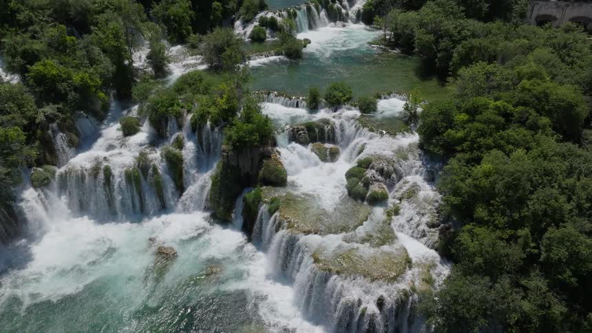 A majestic aerial shot slowly ascends over the powerful, cascading waterfalls of Skradinski Buk in a lush national park