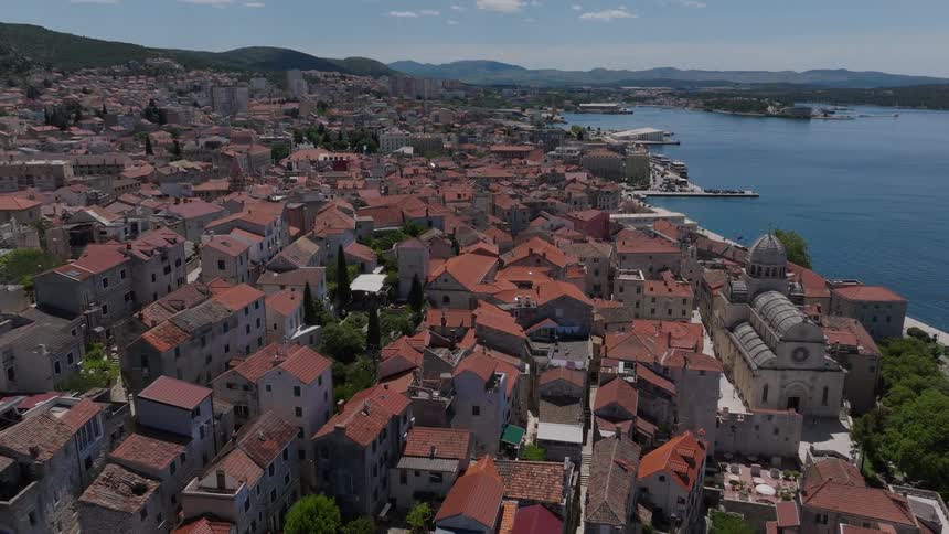 A slow aerial push-in shot reveals the beautiful historic old town of Šibenik, Croatia, with its iconic red-tiled roofs along the sunlit Adriatic coast
