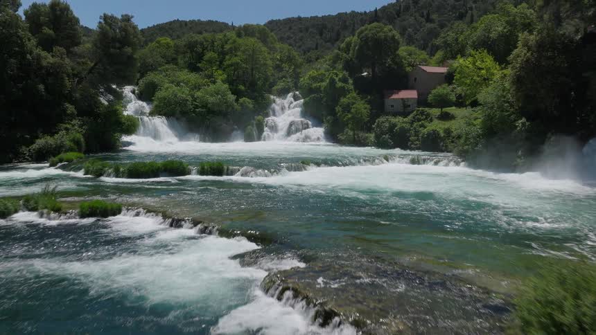 A spectacular aerial view tracks forward over a cascading, multi-tiered waterfall in a lush, green forest