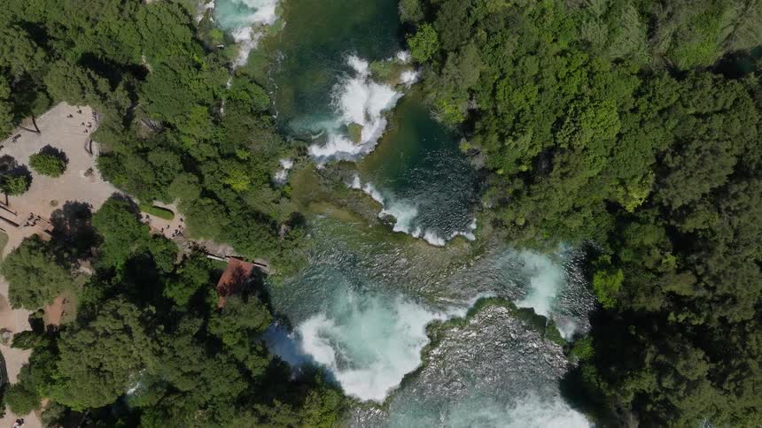 An ascending aerial top-down view reveals a powerful series of cascading waterfalls and turquoise pools surrounded by a lush green forest park