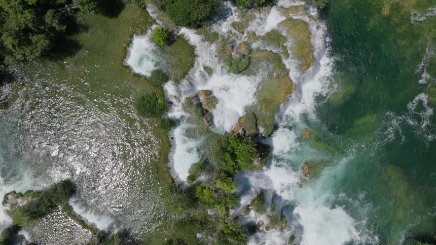 An aerial drone shot slowly rises above a beautiful, wide waterfall cascading through a lush green forest on a sunny day