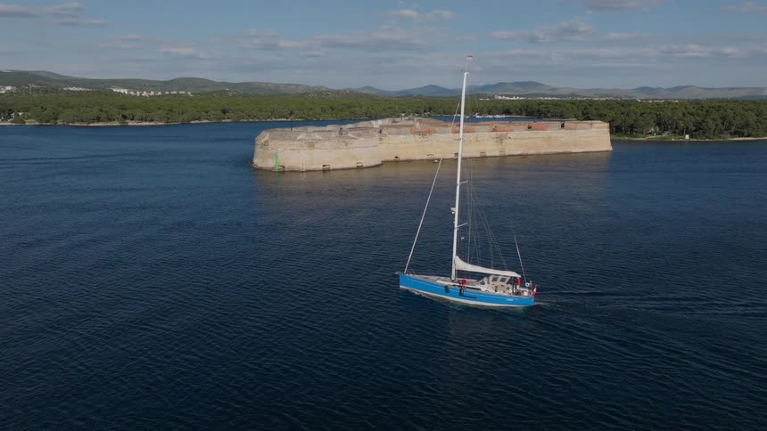 Drone footage captures a blue sailboat gliding through the calm sea past the historic St. Nicholas Fortress on a sunny day