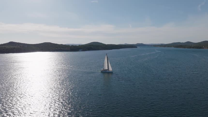 Aerial view of a white sailboat cruising through the calm, shimmering sea on a bright sunny day, surrounded by lush green islands