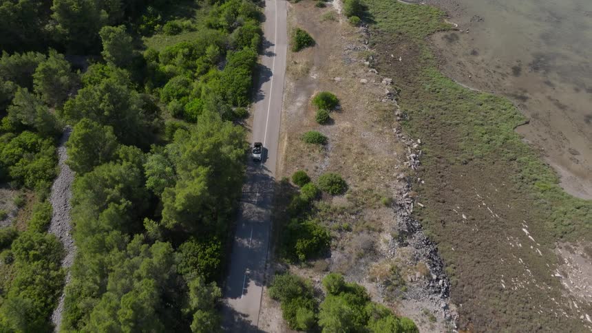 A high-angle aerial view follows a convertible car driving along a scenic coastal road between a lush green forest and the sea