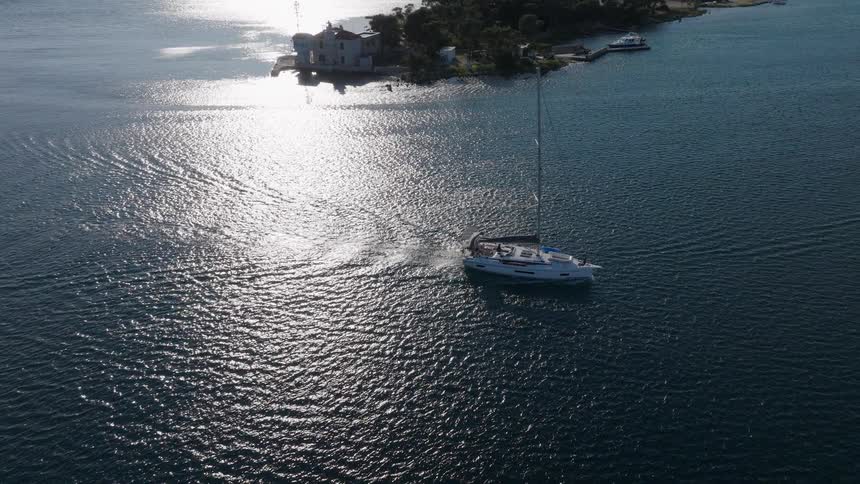 An aerial view follows a sailboat cruising across the shimmering blue sea near a tree-lined island coast on a sunny day