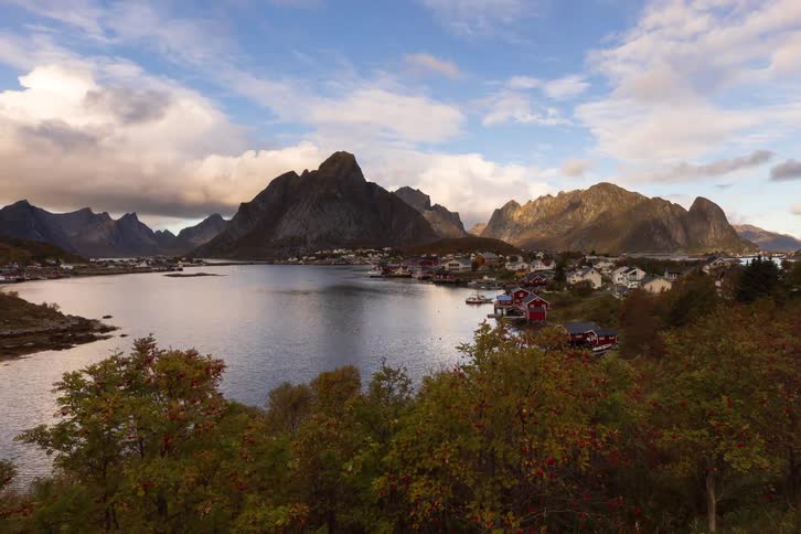 A slow tracking shot reveals the breathtaking autumnal beauty of a Norwegian village nestled in a fjord, surrounded by majestic mountains under a cloudy sky