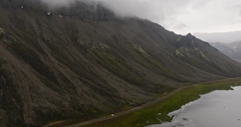 An aerial shot follows a lone white car driving on a remote road between a vast, moody mountain and a tranquil lake
