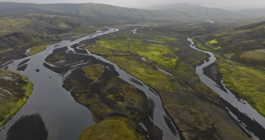 An epic aerial view reveals a vast braided river carving its way through a volcanic landscape with black sand and green moss in Iceland