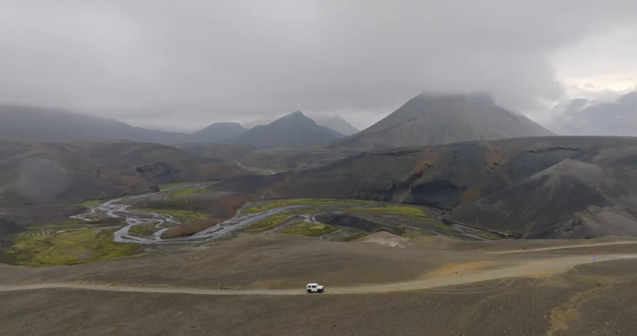 An aerial shot pans across a vast, volcanic landscape in the Icelandic highlands, revealing a lone white SUV parked on a remote road under a foggy sky