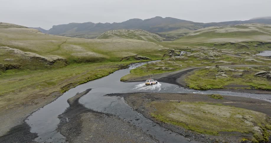 Static aerial view of an adventurous off-road truck driving through a river in the rugged, green landscape of Iceland