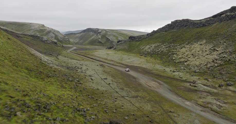 An aerial view flies over a mossy ridge to reveal a lone car on a remote road trip through a vast volcanic valley in Iceland