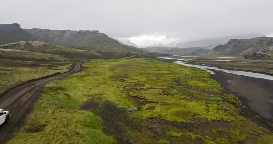 A white off-road vehicle drives along a dirt track through a vast, mossy green valley next to a river in the remote Icelandic highlands