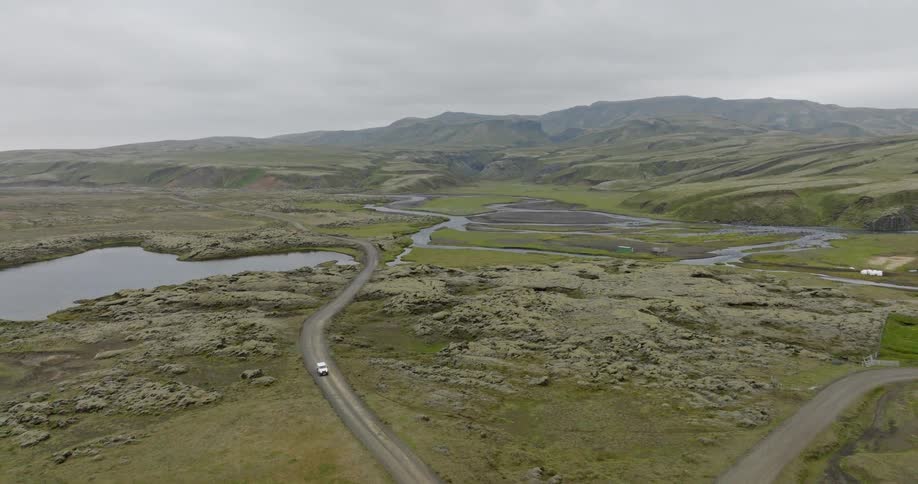 A drone pulls back to reveal a white car driving on a remote road through the vast, mossy volcanic landscape of the Icelandic highlands