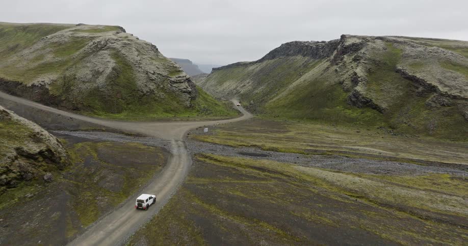 Aerial view of a white SUV driving on a winding dirt road through a vast, mossy green valley in the Icelandic highlands
