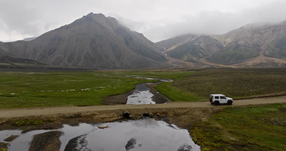 Aerial shot of a white SUV driving through a stunning, remote Icelandic valley with majestic mountains and a winding river