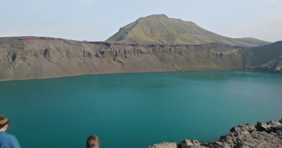 An aerial view pulls back to reveal two hikers standing on the edge of a volcanic crater, overlooking a stunning turquoise lake