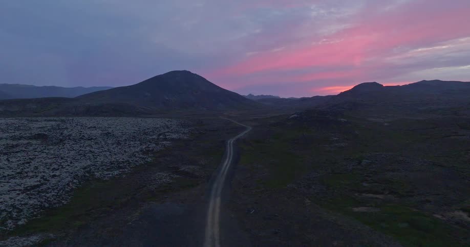 An aerial view captures a winding road through a vast, rocky landscape leading towards distant mountains under a beautiful pink and purple twilight sky