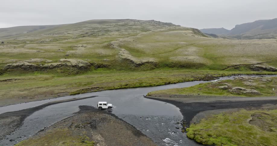 Aerial view of a white 4x4 vehicle driving through a river crossing in the vast, mossy green highlands of Iceland