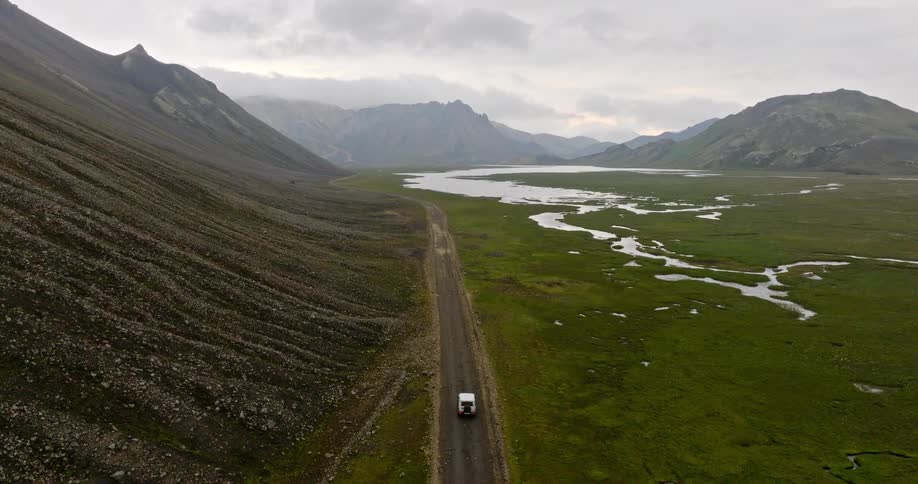 Aerial drone shot reveals a white 4x4 vehicle driving on a remote dirt road through a majestic green valley surrounded by mountains