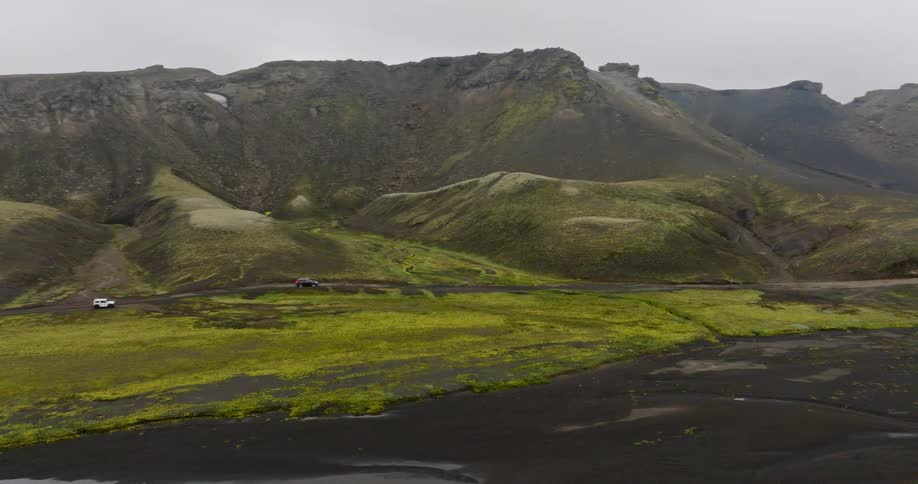 An aerial view reveals a convoy of off-road vehicles navigating a rugged dirt path through the vast, moss-covered volcanic highlands of Iceland