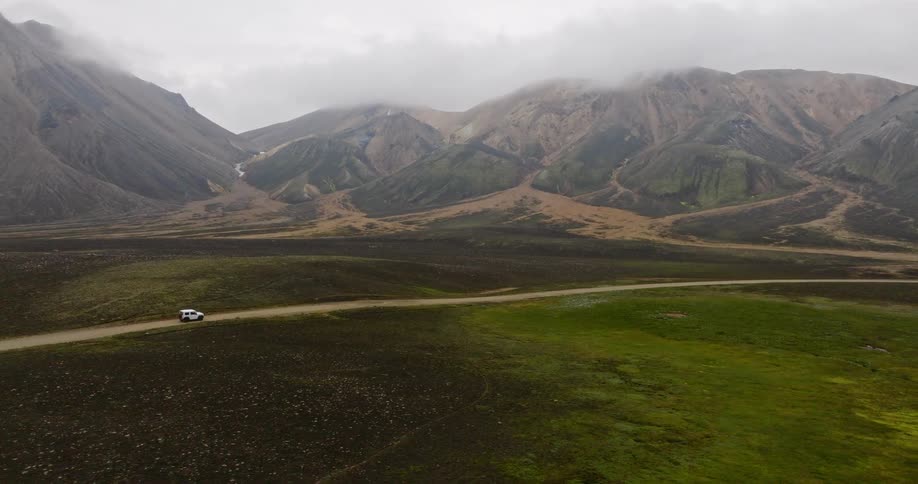 An aerial pullback shot reveals a lone white SUV on a remote dirt road in a vast valley with dramatic, cloud-covered mountains in the background