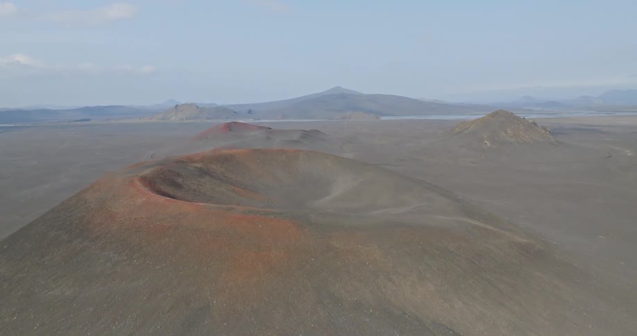 A slow ascending aerial shot reveals a vibrant red volcanic crater in a desolate, barren landscape under a hazy sky