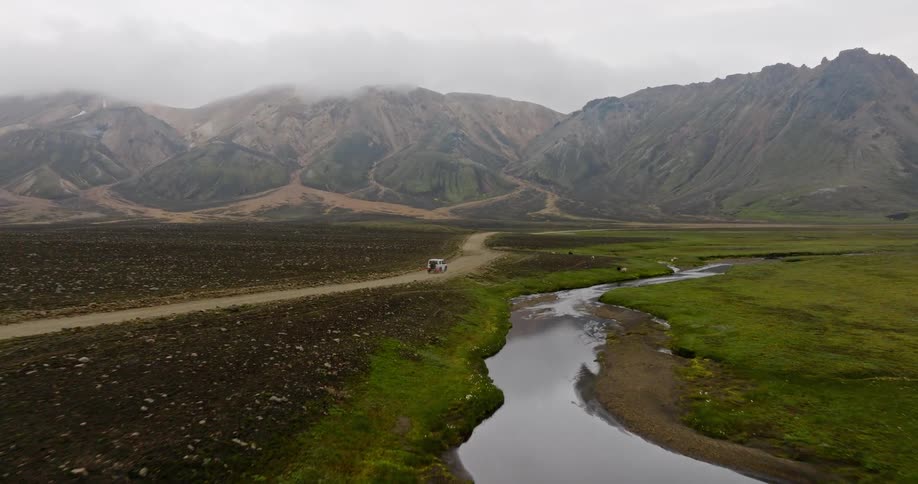 An aerial view captures a white off-road vehicle driving on a remote dirt road through a vast mountain valley with a winding river