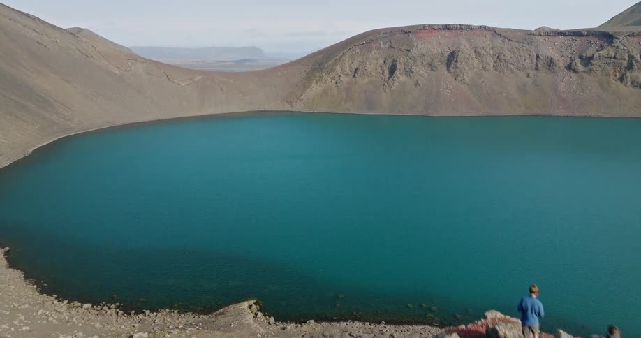 An aerial drone flies past two adventurous hikers standing on the edge of a volcanic crater to reveal a beautiful turquoise lake in Iceland