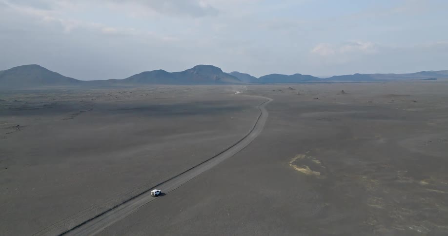 Aerial view of a white car driving along a remote road through a vast, barren volcanic landscape towards distant mountains