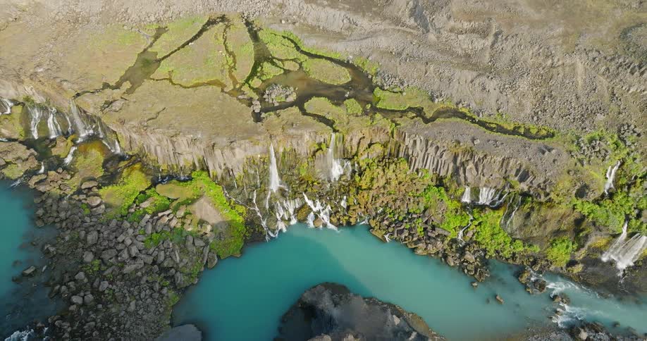An aerial top-down view slowly ascends, revealing a series of stunning waterfalls cascading over a rocky cliff into a vibrant turquoise river