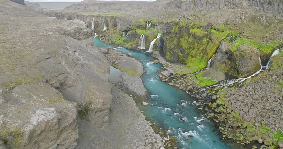 A stunning aerial reveal of a rugged Icelandic canyon where multiple waterfalls flow into a vibrant turquoise river