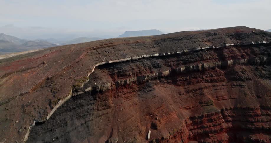 An aerial shot flies over a red and black volcanic crater revealing a vast, desolate river valley in the Icelandic highlands