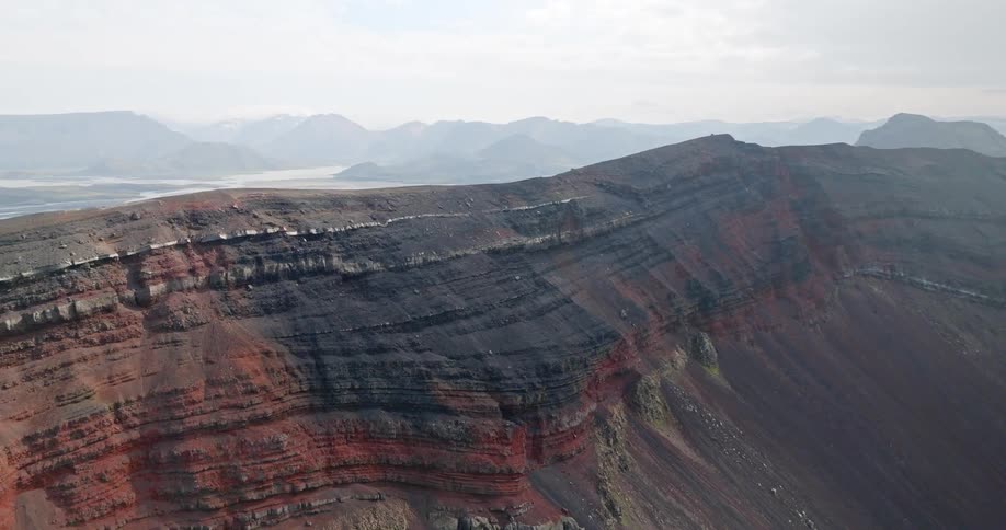A slow aerial pan reveals the colorful, layered rock formations of a massive volcanic crater surrounding a deep blue lake on a misty day