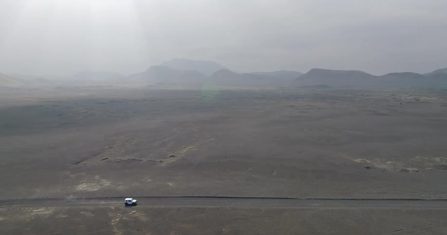 A white SUV drives through a vast, desolate, and foggy volcanic landscape with mountains in the distance