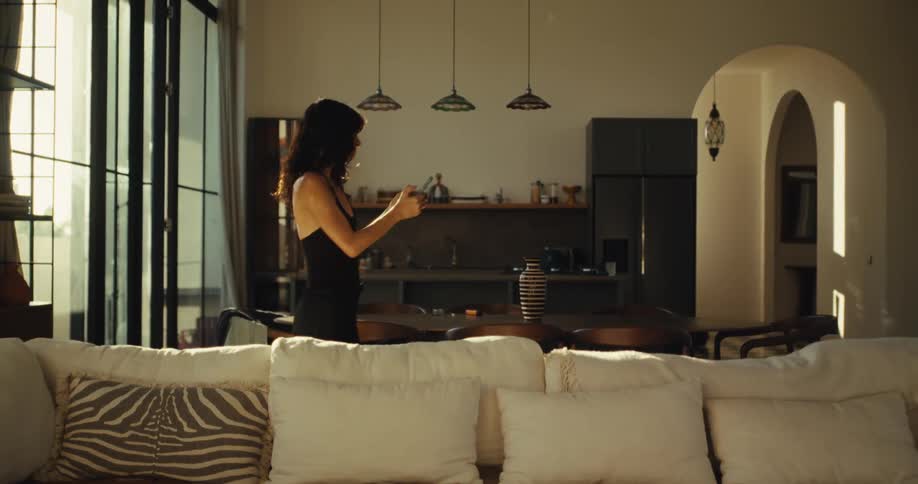 Carefree woman in a sleek black dress grooves to music on her phone in a sun-drenched, modern apartment
