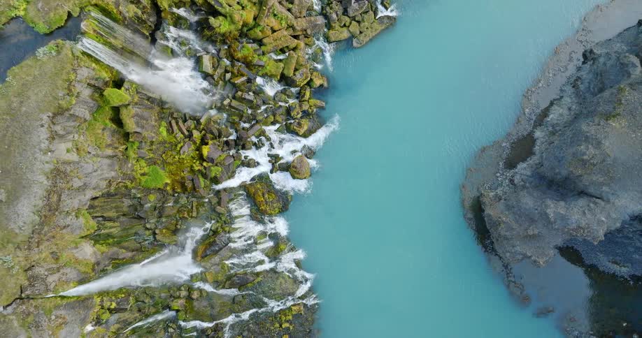 Top-down aerial view slowly ascends to reveal a stunning waterfall cascading over mossy rocks into a vibrant turquoise river