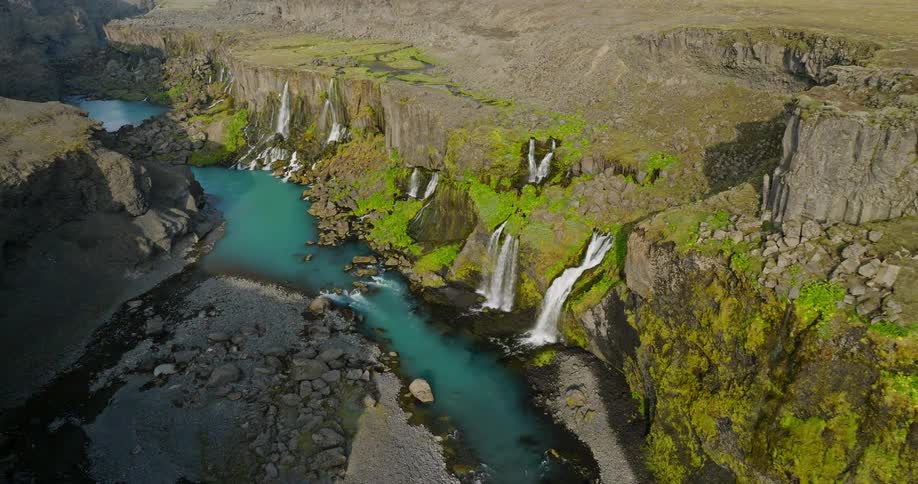 An aerial view rises to reveal a series of stunning waterfalls flowing into a turquoise river within a vast, rocky canyon in Iceland