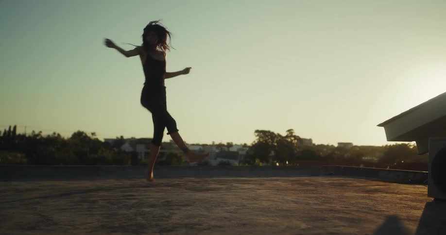A silhouetted woman dances joyfully and expressively on a city rooftop during a golden sunset in slow motion