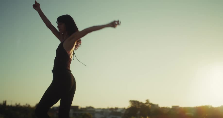 In slow motion, a woman dances freely and barefoot on a rooftop, silhouetted against the warm glow of the setting sun