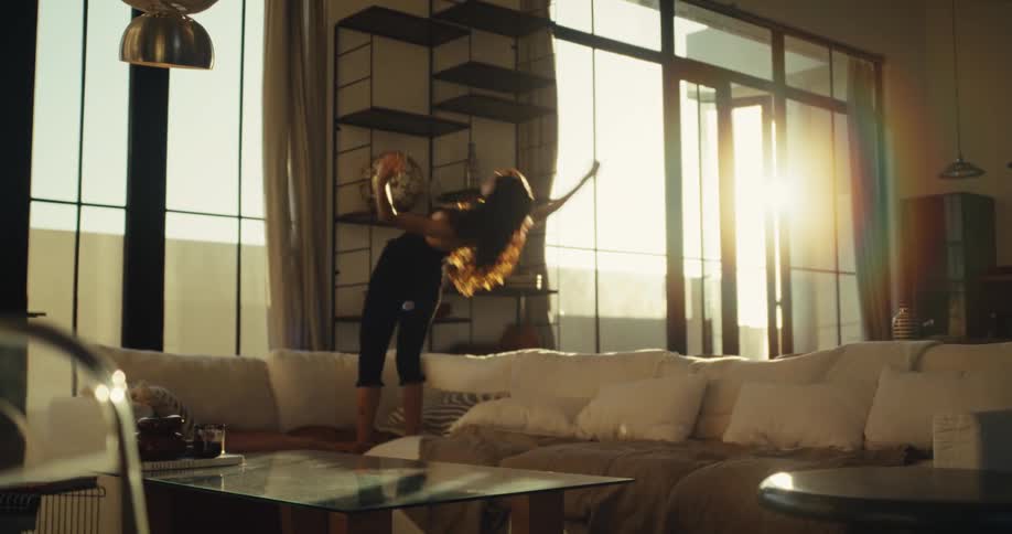 An expressive woman dances with abandon on her couch in a sunlit modern apartment before falling back to rest