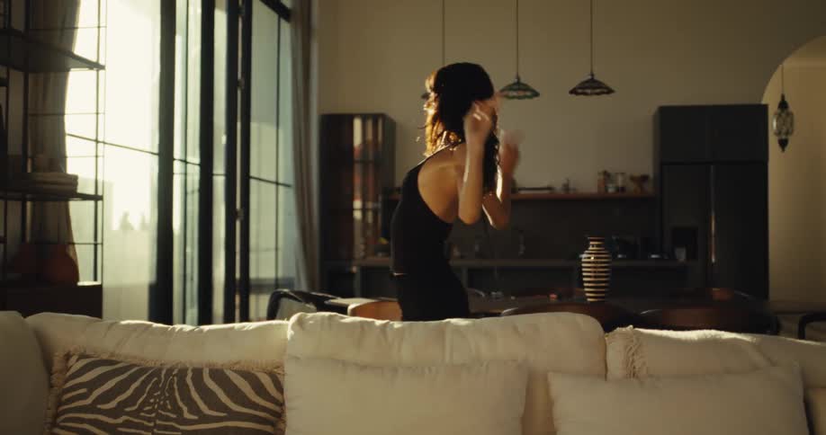 A happy woman dances freely on the back of a sofa in her sun-drenched modern apartment