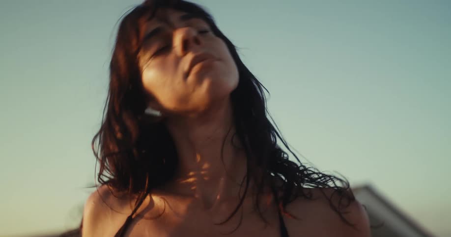 A serene woman with earbuds closes her eyes and moves slowly, her hair blowing in the wind on a rooftop during a beautiful golden hour sunset